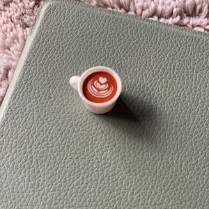 Pode incluir: Uma pequena x&iacute;cara de caf&eacute; branca, cheia de l&iacute;quido marrom e arte latte, colocada sobre uma superf&iacute;cie texturizada verde claro. A x&iacute;cara tem uma pequena al&ccedil;a e cont&eacute;m uma bebida de caf&eacute;. O fundo &eacute; um tecido macio e texturizado.