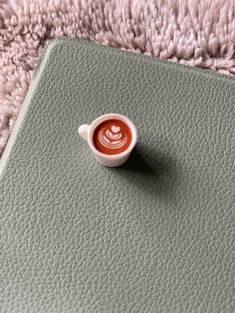 Pode incluir: Uma pequena x&iacute;cara de caf&eacute; branca, cheia de l&iacute;quido marrom e arte latte, colocada sobre uma superf&iacute;cie texturizada verde claro. A x&iacute;cara tem uma pequena al&ccedil;a e cont&eacute;m uma bebida de caf&eacute;. O fundo &eacute; um tecido macio e texturizado.