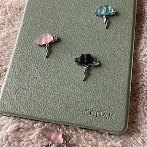 May include: Cloud-shaped pins in pink, blue, and black with silver lightning bolt accents. The pins are on a textured, light green surface with the word "COBAK" visible. One pink pin is separate from the others.