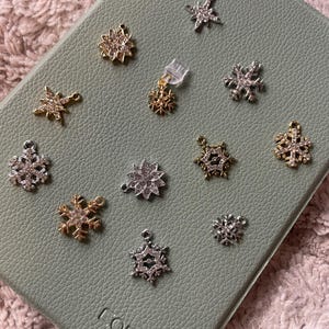 May include: A collection of snowflake charms in gold, silver, and clear crystal. The charms are displayed on a light green leather-like surface. Some charms are dangling, while others are loose. The word "COBAK" is visible on the surface.
