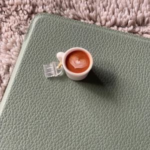 Pode incluir: Uma pequena ch&aacute;vena de caf&eacute; branca com um desenho em forma de cora&ccedil;&atilde;o na espuma, pousada numa superf&iacute;cie verde com textura de couro. A ch&aacute;vena est&aacute; presa a um peda&ccedil;o de pl&aacute;stico transparente com um fecho dourado.
