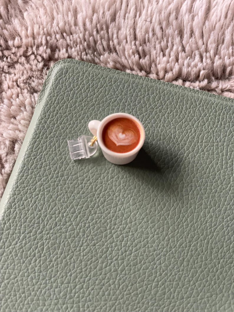 Pode incluir: Uma pequena ch&aacute;vena de caf&eacute; branca com um desenho em forma de cora&ccedil;&atilde;o na espuma, pousada numa superf&iacute;cie verde com textura de couro. A ch&aacute;vena est&aacute; presa a um peda&ccedil;o de pl&aacute;stico transparente com um fecho dourado.