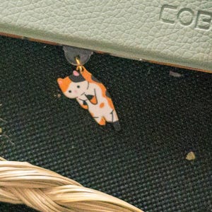 Könnte beinhalten: Ein schwarzer, orangefarbener und weißer Katzenanhänger mit einem goldenen Ring. Der Anhänger hängt an einem Buch.