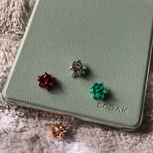May include: An assortment of small, colorful bow charms in red, silver, green, and gold. They are arranged on a light green textured surface, with the word "COBAK" visible. The charms are on a soft, textured background.