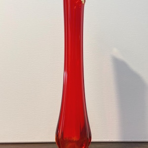 May include: A tall, slender, red glass vase with a ruffled, cream-coloured rim. The vase has a flared base with a gold-coloured, scalloped edge. The vase is approximately 30 cm tall.