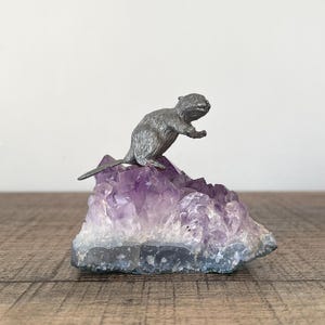 Könnte beinhalten: Eine silberfarbene Biberfigur thront auf einem leuchtend violetten Amethyst-Kristallcluster. Der Kristall hat einen Farbverlauf von tiefem Lila bis Weiß. Die Figur ist detailliert und realistisch.