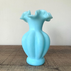 Peut inclure: Un vase bleu clair de style vintage avec un bord froncé. Le vase a un corps bulbeux avec des rainures verticales et une base ronde. Le vase est en matériau opaque et repose sur une surface en bois. Mesure environ 20 cm de haut.