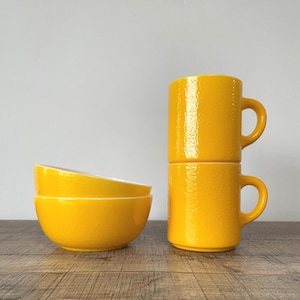May include: Two yellow ceramic mugs stacked, featuring a textured surface and a curved handle. Two yellow bowls are stacked beside the mugs. The items are on a wooden surface.