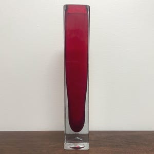 May include: A tall, rectangular glass vase featuring a deep red interior core encased within clear, thick glass walls. The vessel has a flat base and a straight, vertical silhouette, showcasing a smooth, polished surface throughout the entire structure.