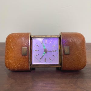 May include: A rectangular travel alarm clock featuring a central square white clock face with gold numerals and hands, encased in a textured tan leather-like folding case with brass accents. The clock face displays the text Europa and 2 Jewels.