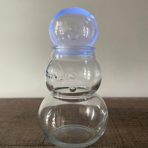May include: A clear glass snowman-shaped jar with a smiling face design. The jar is composed of three rounded sections, with a blue-tinted spherical lid. The jar is sitting on a wooden surface.