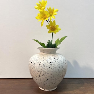 May include: A white vase with a speckled pattern contains a small arrangement of vibrant yellow flowers and green leaves. The vase has a rounded shape and a slender neck. The flowers are in full bloom, providing a splash of colour against the neutral backdrop.