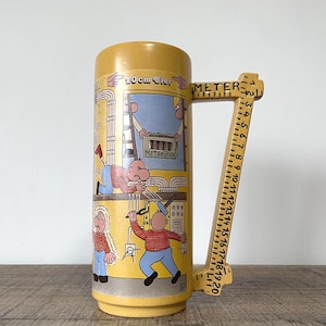 May include: A yellow beer stein with a cartoon illustration of workers and the text "20 cm Bier" and "Meter Bier". The handle is shaped like a ruler with measurements in centimeters.