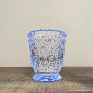 Könnte beinhalten: Ein hellblaues Votivkerzenglas im Vintage-Stil. Das Glas hat ein detailliertes Rautenmuster mit einem gewellten Rand und einem kleinen Sockel. Der Kerzenhalter ist leer und steht auf einer Holzoberfläche.