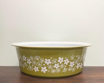 May include: A round, olive green ceramic bowl featuring a white floral pattern around the exterior. The bowl has a wide, flat rim and a smooth, white interior surface. The design consists of various small, stylized flower shapes in white.