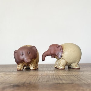 May include: Two ceramic elephant figurines. One elephant is facing forward, the other is facing to the right. Both have brown heads and ears, with beige bodies and legs. The figurines are on a wooden surface.