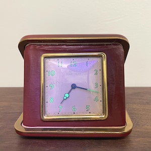 Puede incluir: Un reloj despertador de viaje cuadrado Phinney-Walker con una carcasa de color rojo intenso y un marco de metal dorado. La esfera tiene un fondo claro con números verdes del 1 al 12 y manecillas de hora y minutos con puntas verdes.