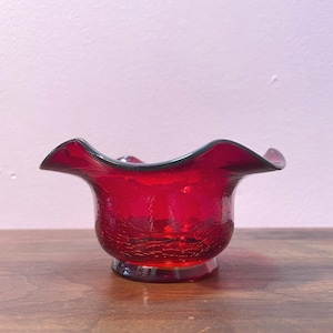 Op de afbeelding: Een robijnrode glazen kom met een gegolfde rand. De kom heeft een craquelé-afwerking en staat op een kleine, ronde basis. Het glas is diep, transparant rood en de rand is iets donkerder.
