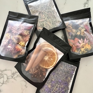 May include: Five black resealable bags filled with various dried herbs and botanicals. Contents include dried flowers, lavender, cinnamon sticks, star anise, and a dried orange slice. The bags are arranged on a white marble surface.