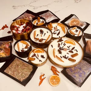 May include: An assortment of handmade candles in various sizes and containers. The candles are white and decorated with dried orange slices, cinnamon sticks, and star anise. Some candles are in gold or black metal containers, while others are in wooden bowls.