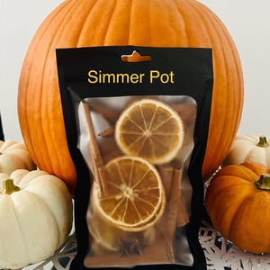 Può includere: Un pacchetto nero con l'etichetta "Simmer Pot" contiene fette d'arancia essiccate, bastoncini di cannella e anice stellato. Il pacchetto è posto su uno sfondo di una grande zucca arancione e diverse zucche bianche e arancioni più piccole.