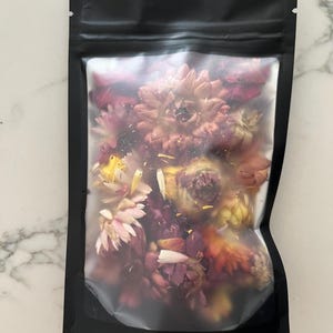 May include: A black, resealable bag filled with dried flowers. The flowers are a mix of pink, red, yellow, and orange hues. The bag is sealed at the top and sits on a white marble surface.