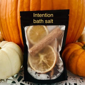 Può includere: Un sacchetto nero con l'etichetta "Intention bath salt" contenente fette d'arancia essiccate, un bastoncino di cannella e sali da bagno. Il sacchetto è posto su uno sfondo di zucche, tra cui una grande zucca arancione e zucche bianche e arancioni più piccole.