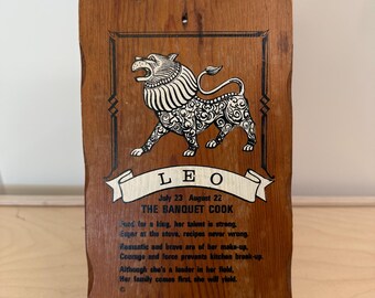 May include: Wooden plaque featuring a detailed illustration of a lion, with the zodiac sign Leo and text "The Banquet Cook." The plaque has a natural wood grain and is supported by a clear plastic stand.