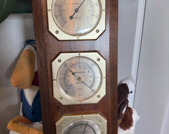 May include: A wooden weather station with three brass-framed gauges. The top gauge reads humidity, the middle one is a barometer, and the bottom one is a thermometer. The wood is a dark brown color.