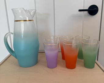 May include: A vintage glassware set featuring a turquoise and white ombre pitcher with a gold rim, and six matching glasses in shades of purple, orange, and green. The glassware is displayed on a wooden tray.