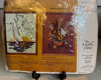 May include: A craft kit package featuring two cross-stitch designs. One depicts a sailboat at sunset, the other a still life with sunflowers. The package includes instructions and materials for each design, with finished sizes of 12" x 16" (30.4 cm x 40.7 cm).