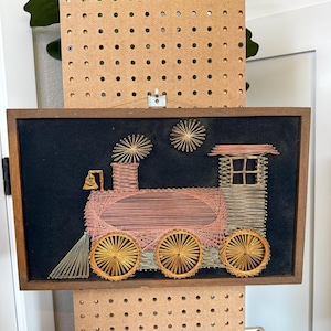 May include: A rectangular wall art piece featuring a string art design of a steam locomotive train. The train is crafted with pink, gold, and silver-toned threads on a black background, enclosed in a simple brown wooden frame with a metal hanger.