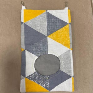 Puede incluir: Una bolsa de almacenamiento rectangular de tela con un patrón geométrico de triángulos amarillos, grises y blancos. La bolsa tiene una abertura circular y clips metálicos en la parte superior para colgar. El diseño es moderno y colorido.