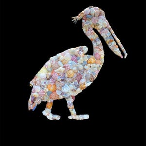 May include: A pelican sculpture constructed from a mosaic of various seashells in shades of white, tan, orange, pink, and brown. The bird is depicted in profile with its long beak slightly open, showcasing the intricate texture of the shells.
