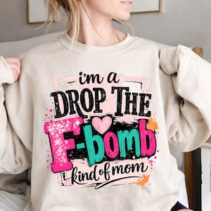Puede incluir: Sudadera beige con el texto "I'm a DROP THE F-BOMB kind of mom." El texto es en negro, rosa y turquesa, con un corazón rosa y salpicaduras de colores. Una prenda audaz y divertida.