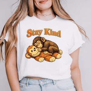 Pode incluir: Camiseta branca com uma ilustração de desenho animado de dois macacos. Um macaco está dormindo em cima do outro. O texto "Stay Kind" é impresso acima dos macacos em letras laranja.