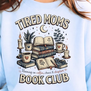 Op de afbeelding: Lichtblauw sweatshirt met een afbeelding van "Tired Moms Book Club". Het ontwerp toont boeken, koffiemokken, planten, kaarsen en de tekst "Running on coffee, chaos & chapters."