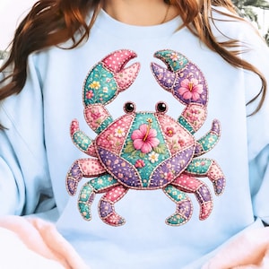 May include: Light blue sweatshirt featuring a colorful crab design. The crab is made of patchwork fabric with pink, teal, and purple sections. It has a pink hibiscus flower in the center and floral accents.