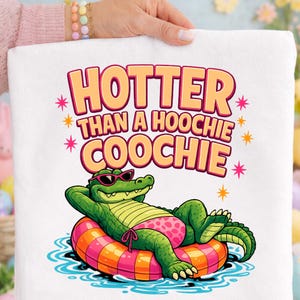 May include: White towel featuring a cartoon alligator wearing sunglasses and a pink swimsuit, lounging on an orange and pink inflatable ring. The towel has the text "HOTTER THAN A HOOCHIE COOCHIE" in orange and pink.