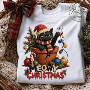 May include: A white t-shirt featuring a graphic of a black cat wearing a red Santa hat and a striped scarf, sitting in a cracked terracotta pot. The cat holds a string of Christmas lights. The text reads Meowy Christmas in red and black.