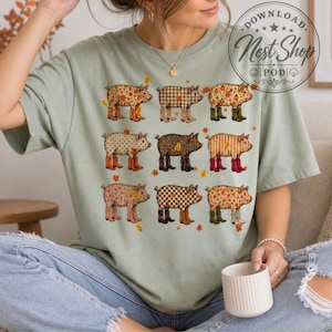 Peut inclure: Un t-shirt vert sauge avec une impression graphique de neuf cochons disposés en grille. Chaque cochon présente un motif différent, notamment à carreaux, léopard et floral, et porte des bottes de pluie colorées parmi des feuilles d'automne.