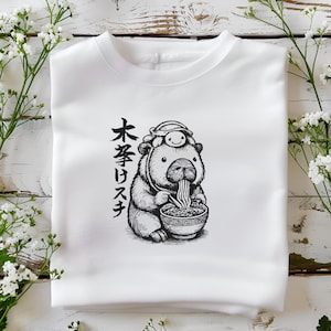 May include: White sweatshirt featuring a black and white illustration of a bear-like creature eating ramen from a bowl. The creature wears a hat and has Japanese text on the left side. The sweatshirt is folded on a white surface.