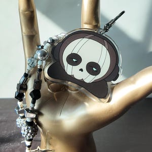 May include: A clear acrylic charm featuring a cartoon skull wearing headphones. The skull has a white face with black eyes and a brown outline. A beaded lanyard with black, silver, and clear beads is attached to the charm.