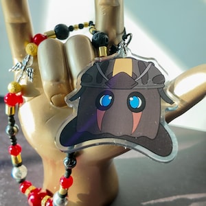 May include: A clear acrylic charm featuring a cartoon character with a dark brown hat and blue eyes. The charm is attached to a beaded bracelet with red, gold, black, and silver beads. The charm is held up by a gold hand.