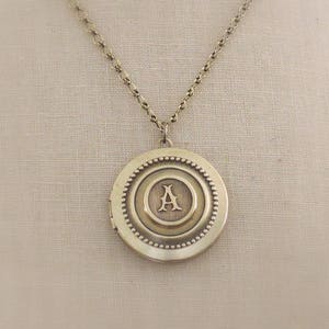 May include: A gold necklace with a round locket pendant featuring the letter "A" in a decorative font.