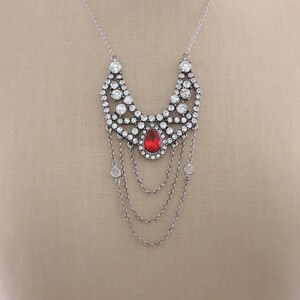May include: A silver necklace with a large, red, teardrop-shaped crystal surrounded by smaller clear crystals. The necklace has a cascading chain design.