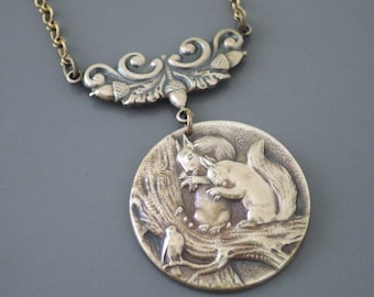 May include: A vintage-style pendant necklace with a detailed relief of squirrels and a bird on a tree branch. The round pendant is suspended from a decorative filigree and a gold-toned chain. The pendant is approximately 1.5 inches in diameter.