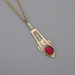 May include: A gold necklace with a pendant featuring a geometric design and a red gemstone.