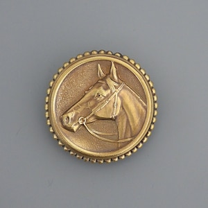 May include: A gold-toned metal pin featuring a detailed, embossed image of a horse's head. The horse is facing left and has a bridle on.