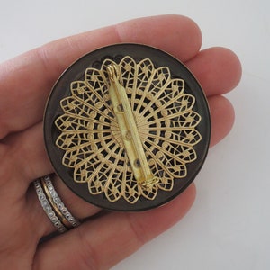 Peut inclure: Une broche ronde noire et dor&eacute;e avec un motif d&eacute;coratif en filigrane. La broche a un fermoir &agrave; &eacute;pingle.